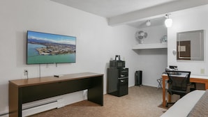 Une chambre d’hôtel comprenant un lit, un bureau avec une chaise, un téléviseur fixé au mur, une petite kitchenette et une fenêtre donnant sur un paysage urbain.