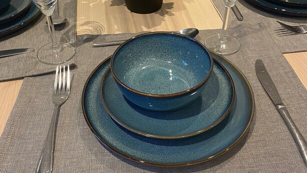 Mesa de jantar redonda posta com pratos azuis, copos transparentes e um vaso com flores.