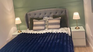 Quarto com cabeceira estofada, edredom azul escuro, duas luminárias de apoio e parede com detalhes verdes.