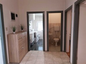 A narrow hallway leading to a bathroom with a toilet, sink, and washing machine. There are two potted plants on a wooden cabinet.