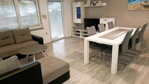 Una sala moderna con un sofá, una mesa redonda de centro y una mesa de comedor con sillas. Se ve un televisor y un mueble en el fondo.