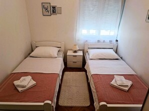 A small, clean room with two single beds, a nightstand, and a window with blinds.
