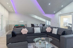 Een moderne woonkamer met een hoekbank, een glazen salontafel en een pooltafel op de achtergrond.