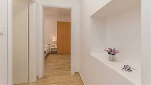 Ein schmaler Gang mit Holzboden führt zu einem Schlafzimmer mit Bett und Nachttisch. Eine weiße Ablagefläche mit einer Blumenvase ist auf der rechten Seite sichtbar.