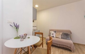 Un petit salon accueillant, avec une table ronde blanche, une chaise en bois, un canapé recouvert d’un plaid et un vase avec des fleurs accroché au mur.