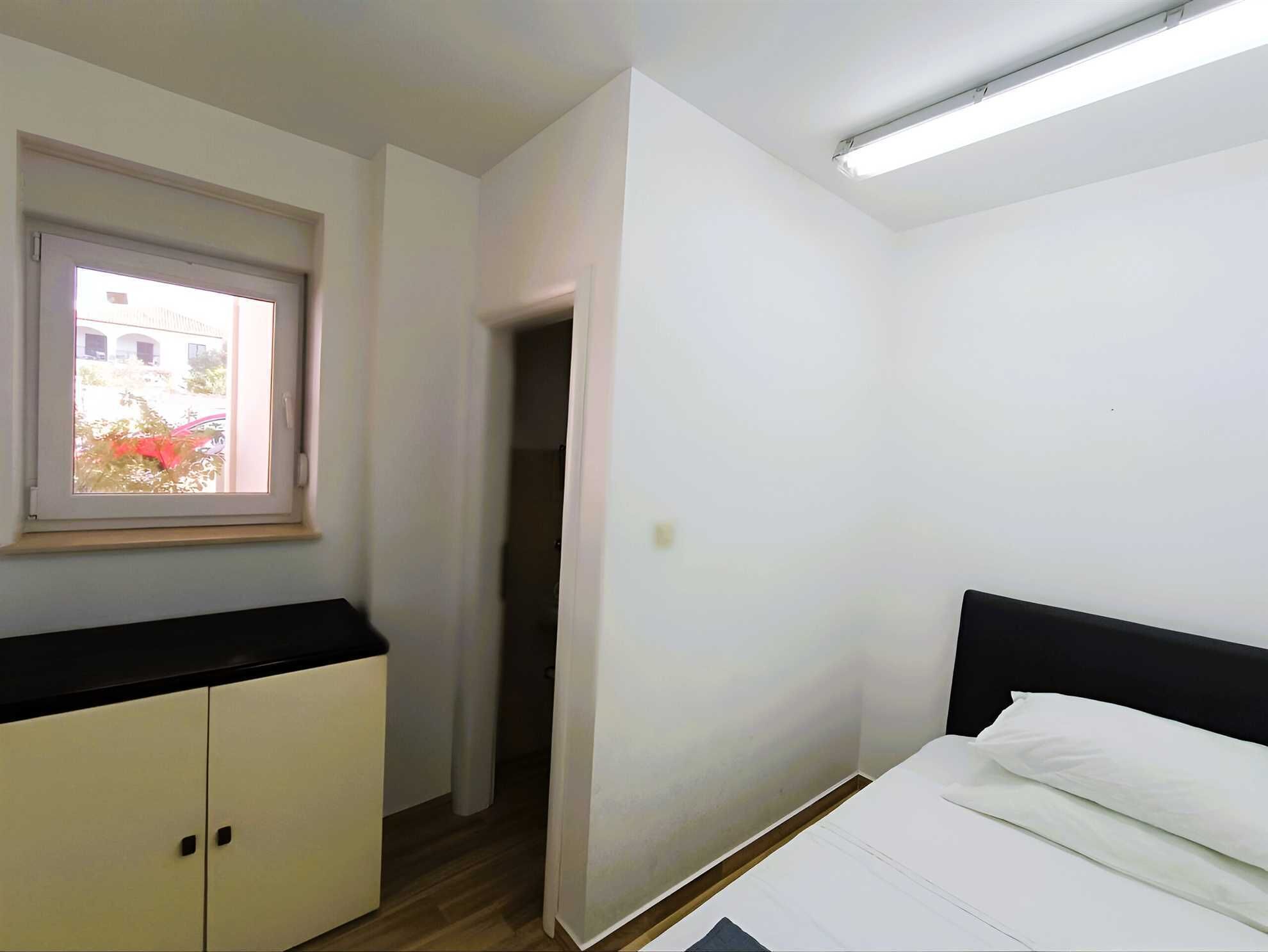 A bedroom with a bed, a window, a wardrobe, and a door leading to another room.