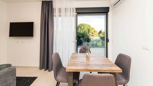 Une pièce moderne avec une table à manger en bois, deux chaises grises, un téléviseur à écran plat fixé au mur et une grande fenêtre donnant sur un paysage verdoyant.