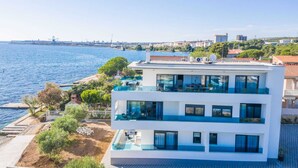 Un immeuble moderne à deux étages, doté d’une piscine sur le toit, situé près du bord de l’eau et offrant une vue sur la mer et le paysage urbain lointain.