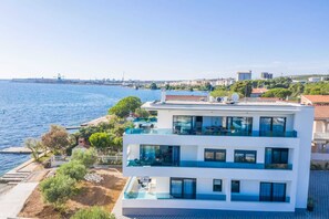 A modern two-story building with a rooftop pool, located near a waterfront with a view of the sea and distant cityscape. - Beachfront Studio Deluxe Apartment Fresca (Bibinje)