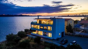 Ein modernes, zweistöckiges Haus mit Balkon in der Nähe von Wasser, mit Blick auf den Sonnenuntergang.