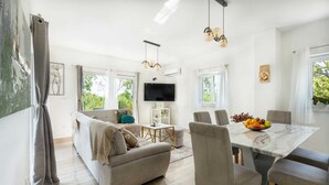 Una sala moderna con sofá, mesa de comedor y sillas, televisor de pantalla plana y ventanas amplias con vistas a zonas verdes.