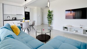 Ein modernes Wohnzimmer mit einem blauen Sofa, einem Essbereich mit Tisch und Stühlen, einer Küche mit weißen Schränken und einem an der Wand befestigten Fernseher.