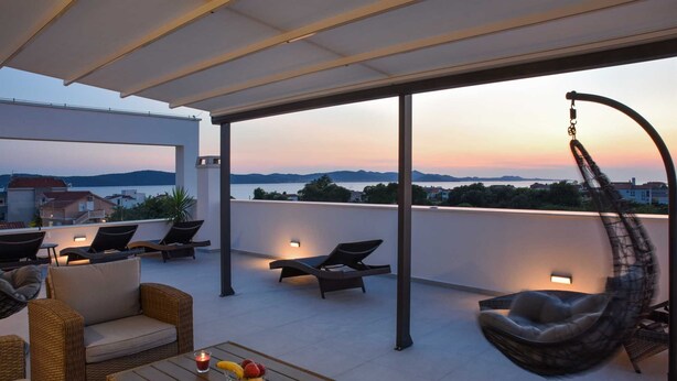 Eine Dachterrasse mit Korbmöbeln, einem Hängesessel und Blick auf das Meer bei Sonnenuntergang.