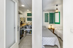 Cocina compacta con lavamanos, estufa y repisas con vajilla. Hay dos camas con ropa de cama estampada y una mesita de noche con lámpara.