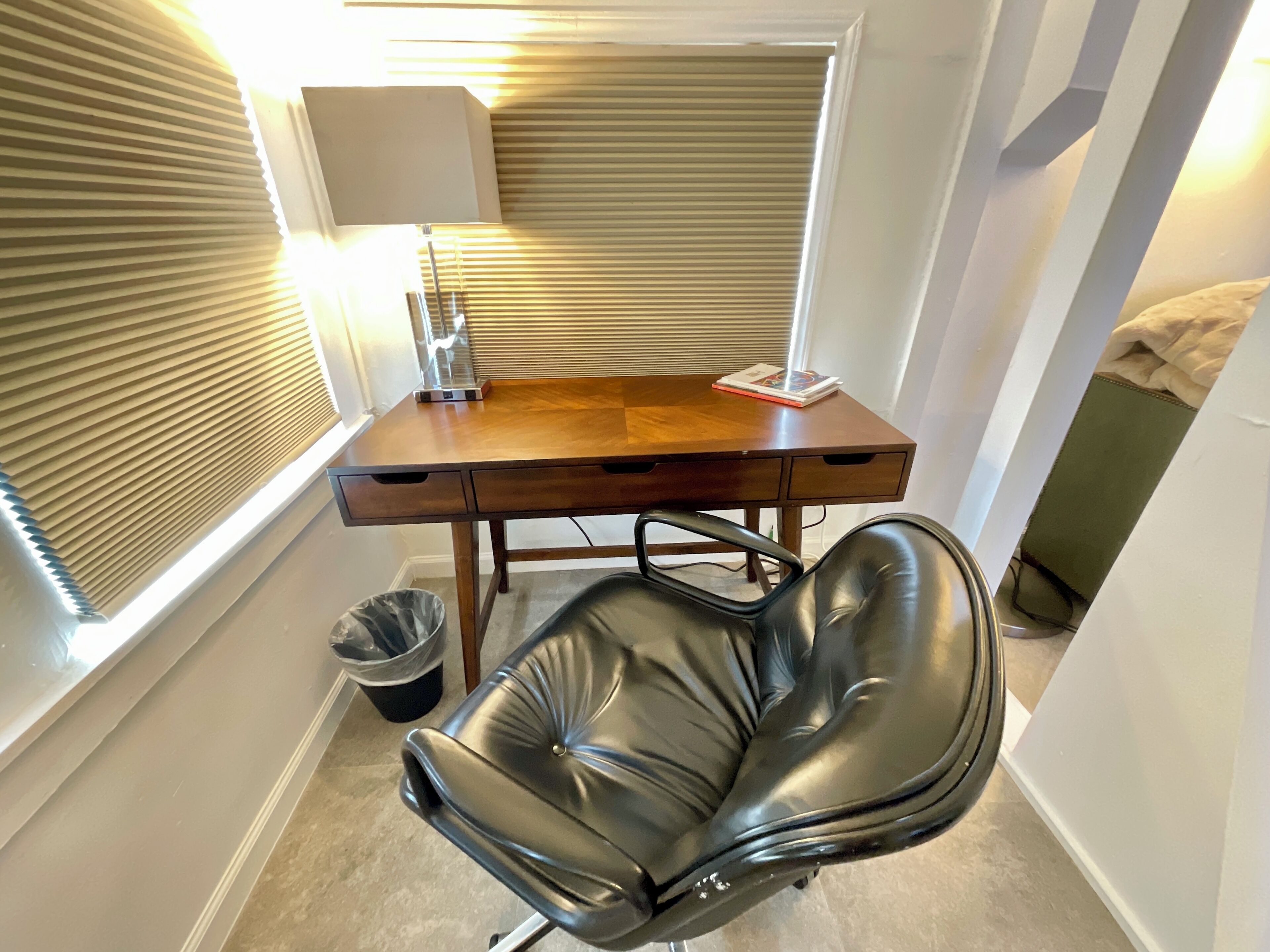 Un petit bureau avec un bureau en bois, une chaise en cuir noir, une lampe et une fenêtre avec des stores.