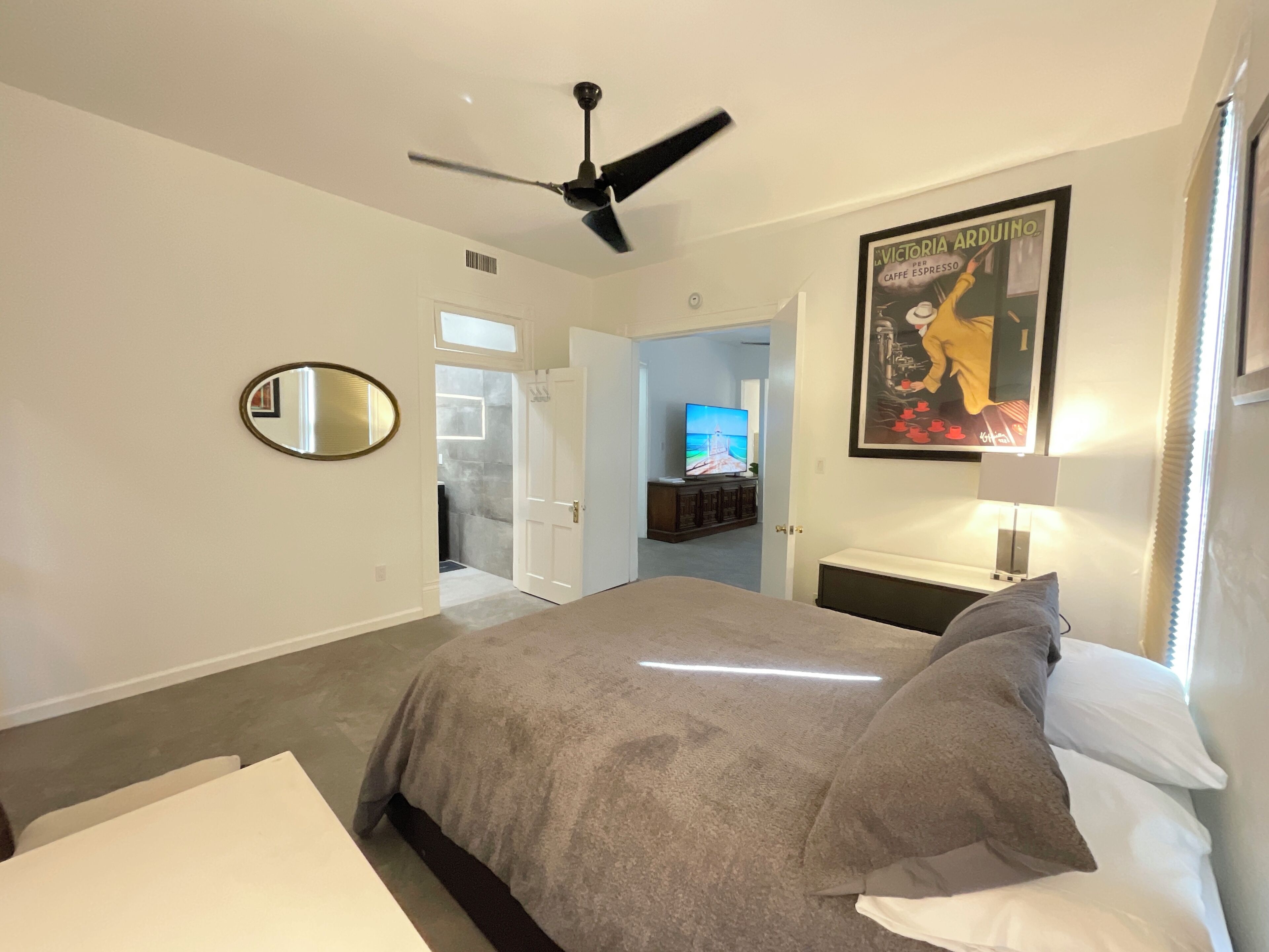 A bedroom with a bed, a ceiling fan, a mirror, a television in the adjacent room, and a poster on the wall.