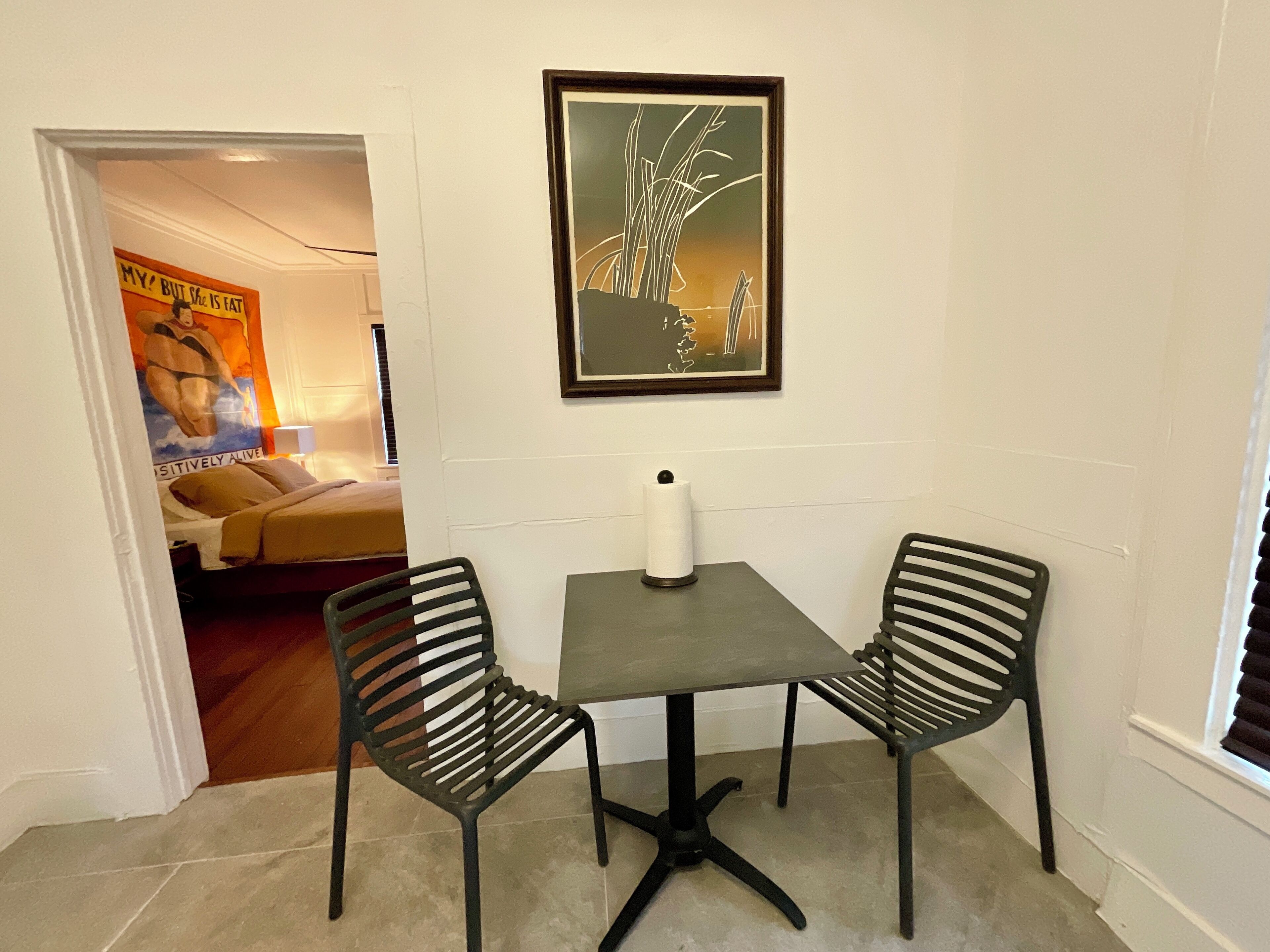 Ein kleiner Raum mit einem Tisch und zwei Stühlen, einem gerahmten Bild an der Wand und einem Plakat an der Tür.