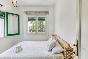 Ein ordentlich bezogenes Bett mit Kissen und einem gefalteten Handtuch, ein Fenster mit grünen Rollläden sowie eine an der Wand befestigte Lampe.