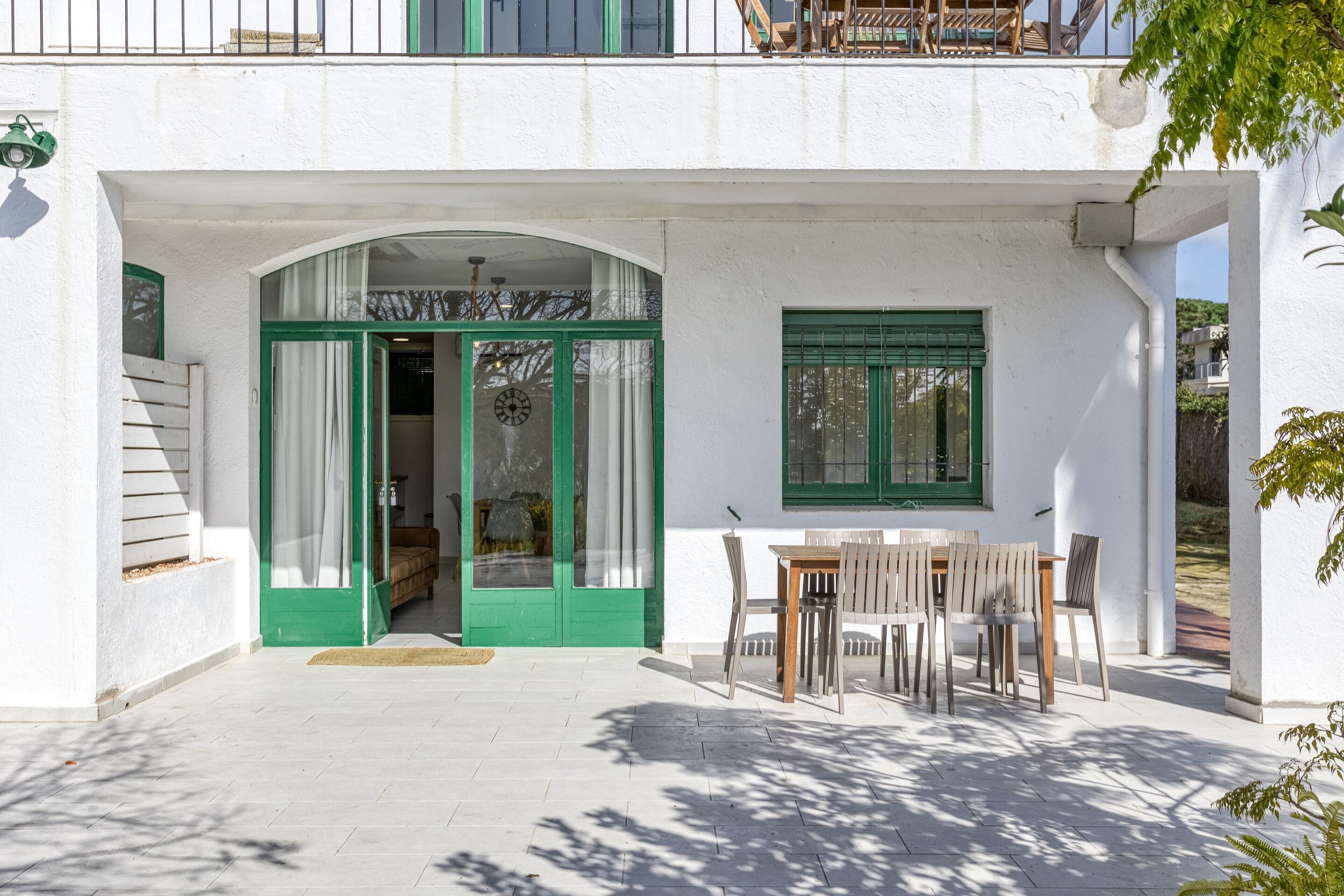 A white building with green-framed doors and windows, a covered patio area with a table and chairs, and a staircase leading to an upper level.