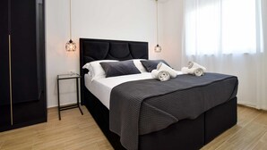A modern bedroom with a black bed, white bedding, and a checkered throw blanket. There are two bedside tables with lamps, a black wardrobe, and a window with sheer curtains. - Apartment with rooftop pool A2, Bibinje (Bibinje)
