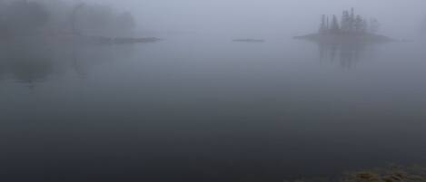Um lago enevoado com margem rochosa e vegetação escassa.