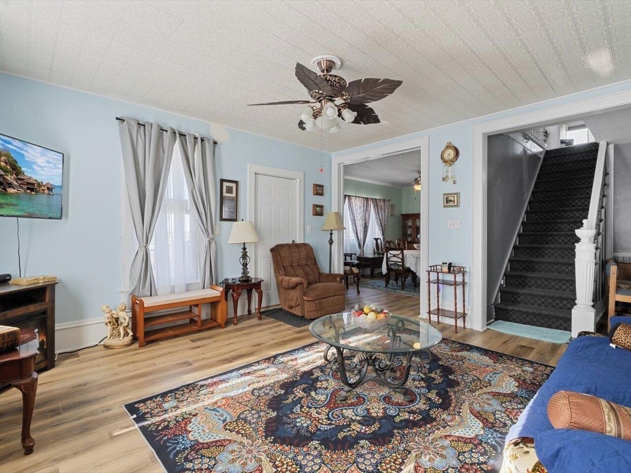 A living room with a wooden floor, a glass coffee table, a brown armchair, a blue sofa, a patterned rug, a ceiling fan, and a staircase leading to another room.