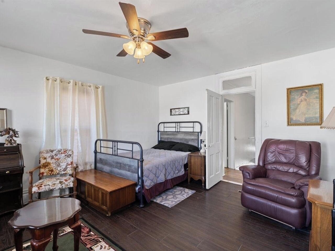Une chambre à coucher comprenant un lit, une chaise, un bureau, un ventilateur de plafond et une porte donnant sur une autre pièce.
