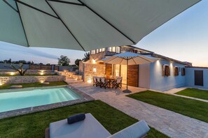Un patio moderno al aire libre con piscina, muros de piedra y un área de comedor con sombrillas.