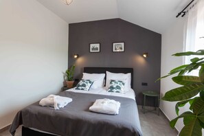 Un dormitorio moderno con un cabecero oscuro, dos luces fijadas a la pared, una planta grande en maceta y una mesita de noche pequeña.