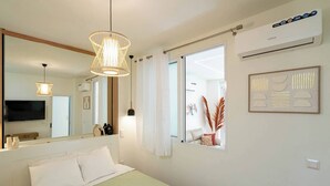Une chambre d’hôtel comprenant un lit, un grand miroir, un luminaire au plafond, une fenêtre avec des rideaux et un climatiseur fixé au mur.