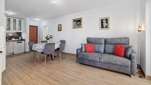 Een woonkamer met een grijze bank, een eettafel met stoelen, een keuken en een witte muur met ingelijste foto's.