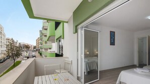 Een balkon met een tafel en stoelen, een schuifdeur naar een slaapkamer, en uitzicht op de straat en gebouwen.