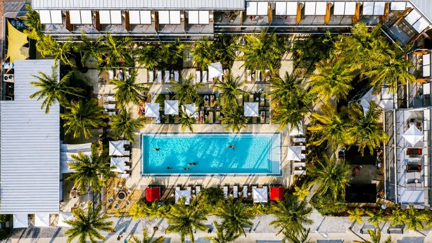 An aerial view of a resort with a central swimming pool surrounded by palm trees and sun loungers.