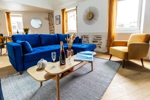 A living room with a blue sofa, a wooden coffee table with a book and glasses, a yellow armchair, and a stone wall. - Grand duplex 100 m² downtown Quimper - 3 bedrooms - All on foot (Quimper)