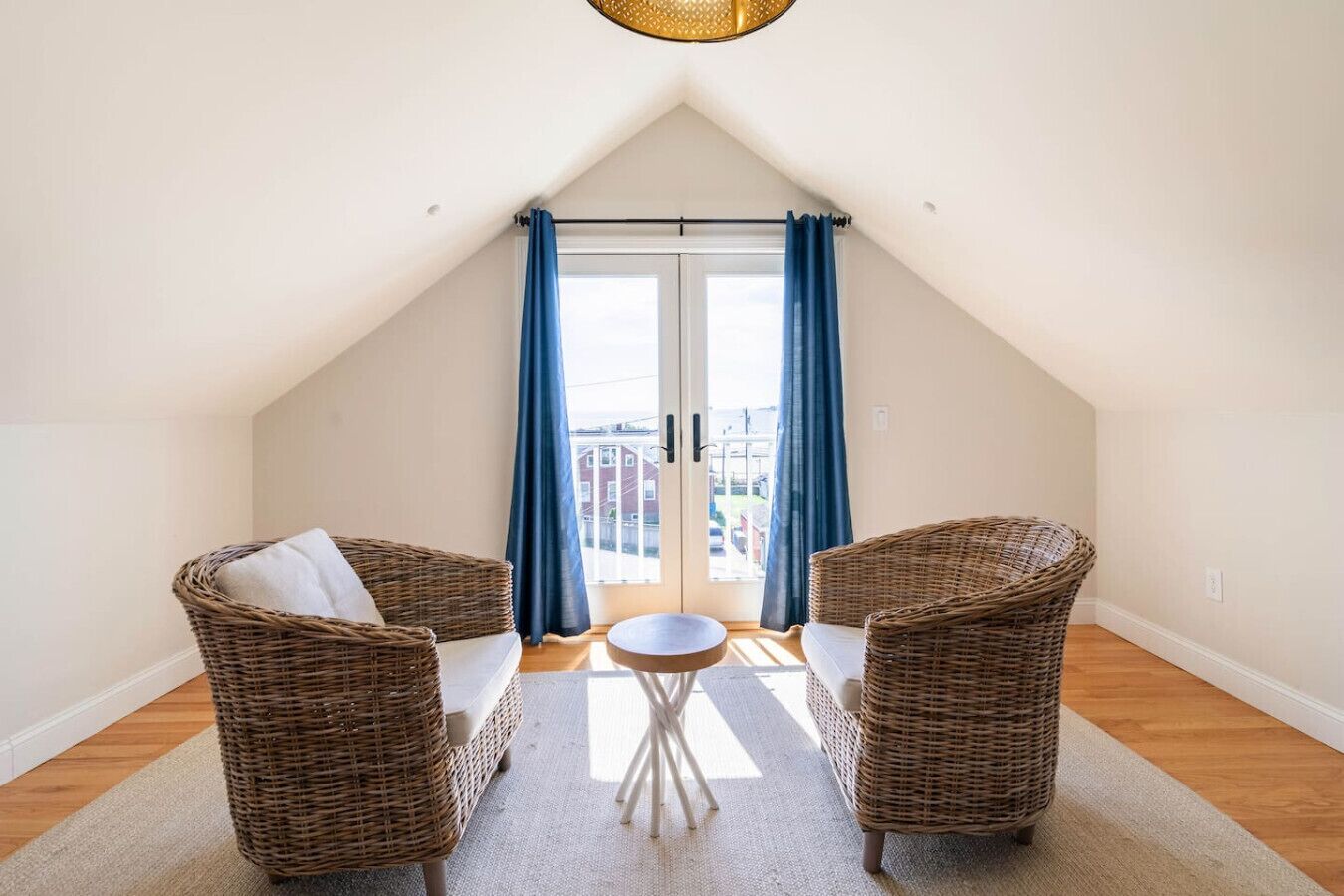 Une pièce chaleureuse avec un plafond en pente, deux fauteuils en osier, une petite table ronde et une vue sur l’extérieur, visible à travers une porte vitrée ornée de rideaux bleus.