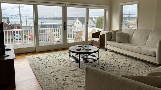A living room with a sofa, a coffee table, and a TV stand. There are large windows with a view of a body of water and buildings.