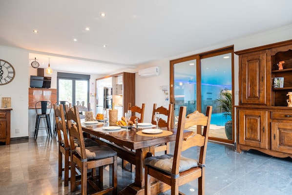 Un coin repas avec une table et des chaises en bois, une cuisine à l’arrière-plan et une vue sur une piscine, visible à travers de grandes portes vitrées.
