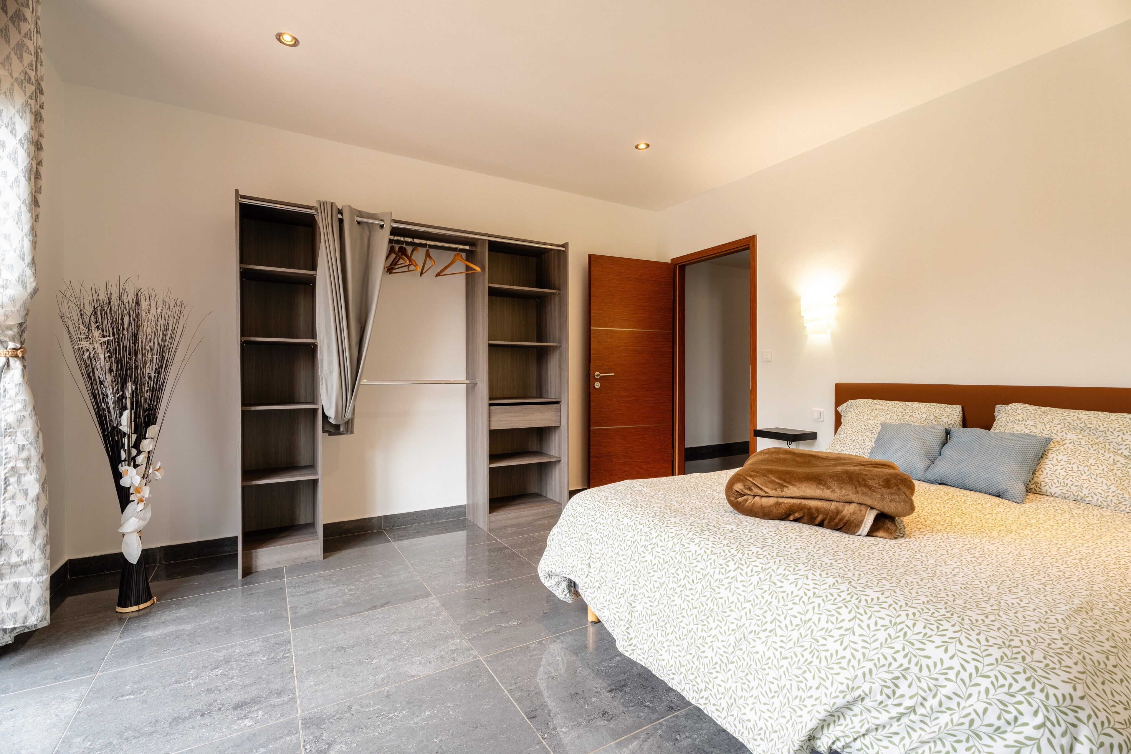 A bedroom with a bed, a wardrobe, a mirror, and a vase with dried plants.