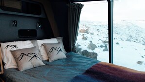 Ein Bett mit Kopfkissen mit Berggermotiv, einer Decke und Blick auf eine verschneite Landschaft durch ein Fenster.