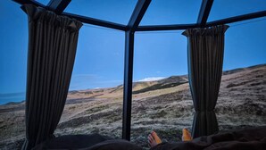 Ein Zimmer mit einem großen Fenster, Vorhängen und Blick auf eine hügelige Landschaft.