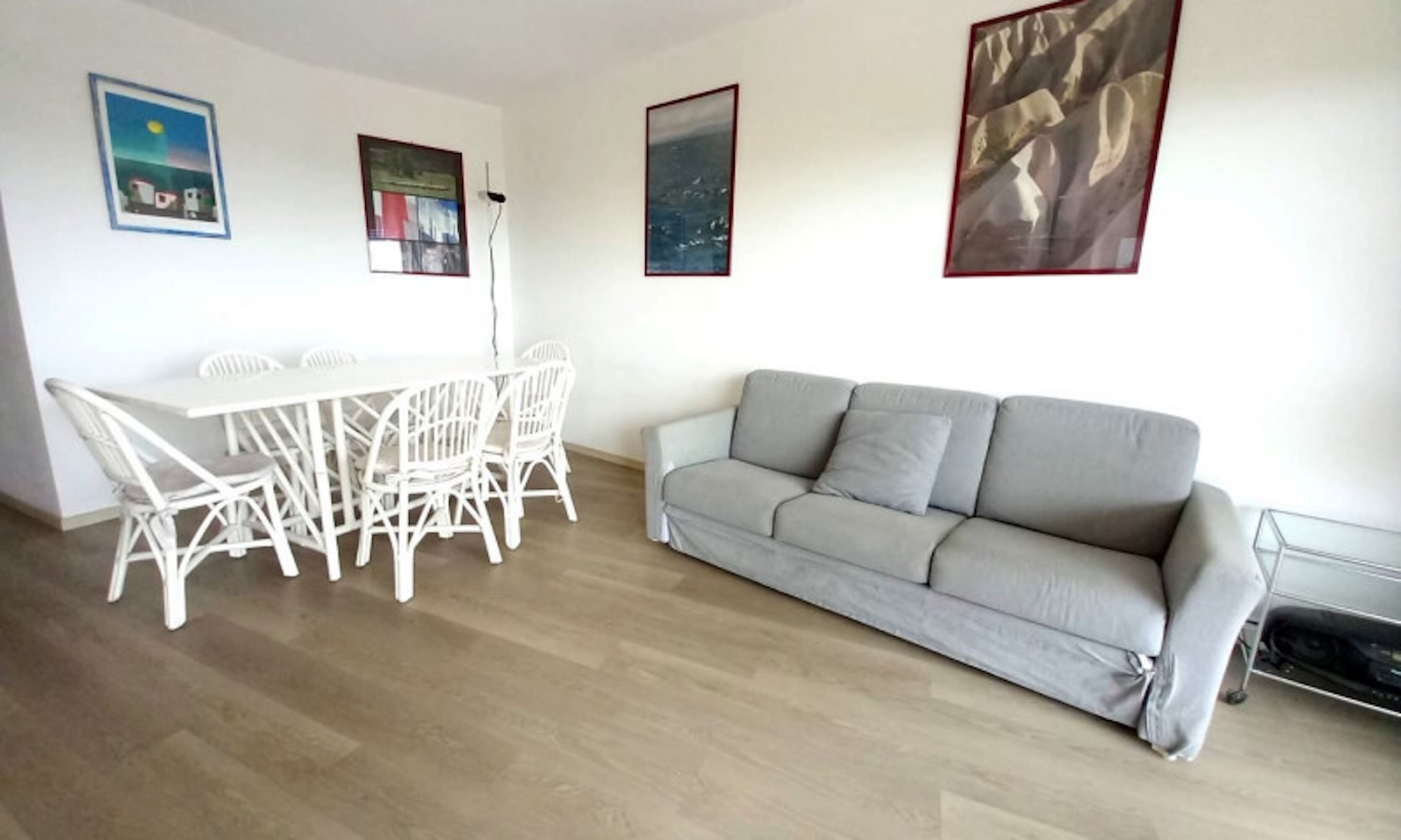 Un salon avec une table et des chaises de salle à manger blanches, un canapé gris et deux œuvres d’art encadrées accrochées au mur.