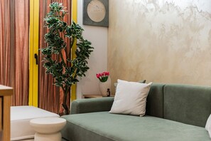 Un canapé vert avec un coussin blanc, une petite table blanche, une grande plante en pot et un vase rempli de fleurs roses.