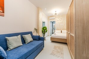 A modern hotel room with a blue sofa, a wooden wardrobe, a bed with white bedding, a small potted plant, and a textured rug. - NooN Properties | 3 by PikHost (Dhërmi)