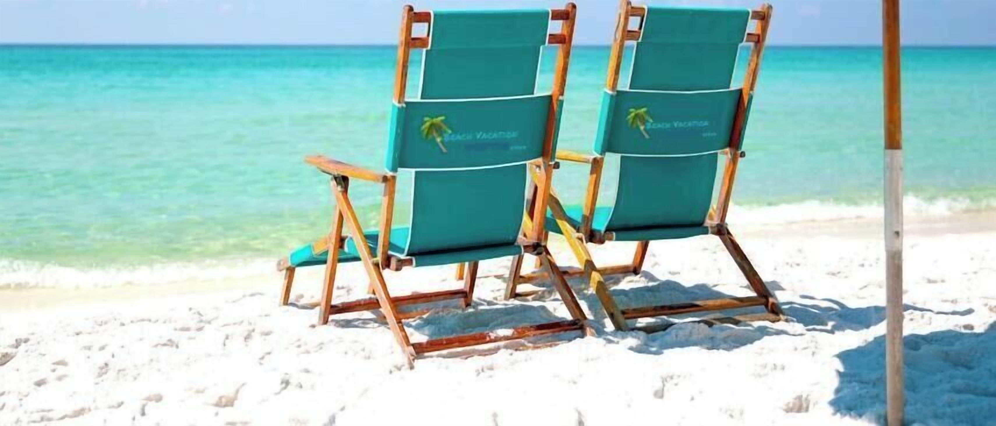 Zwei Strandstühle unter einem großen Sonnenschirm an einem sandigen Strand, im Hintergrund klares türkisblaues Wasser.