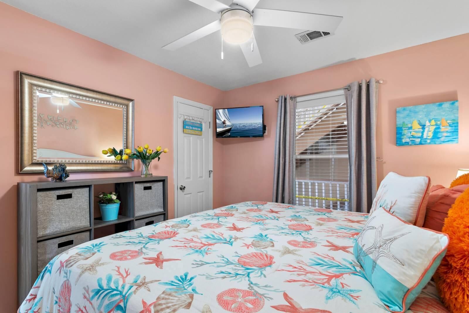 A bedroom with a bed, a dresser, a mirror, a TV, and a window with curtains.