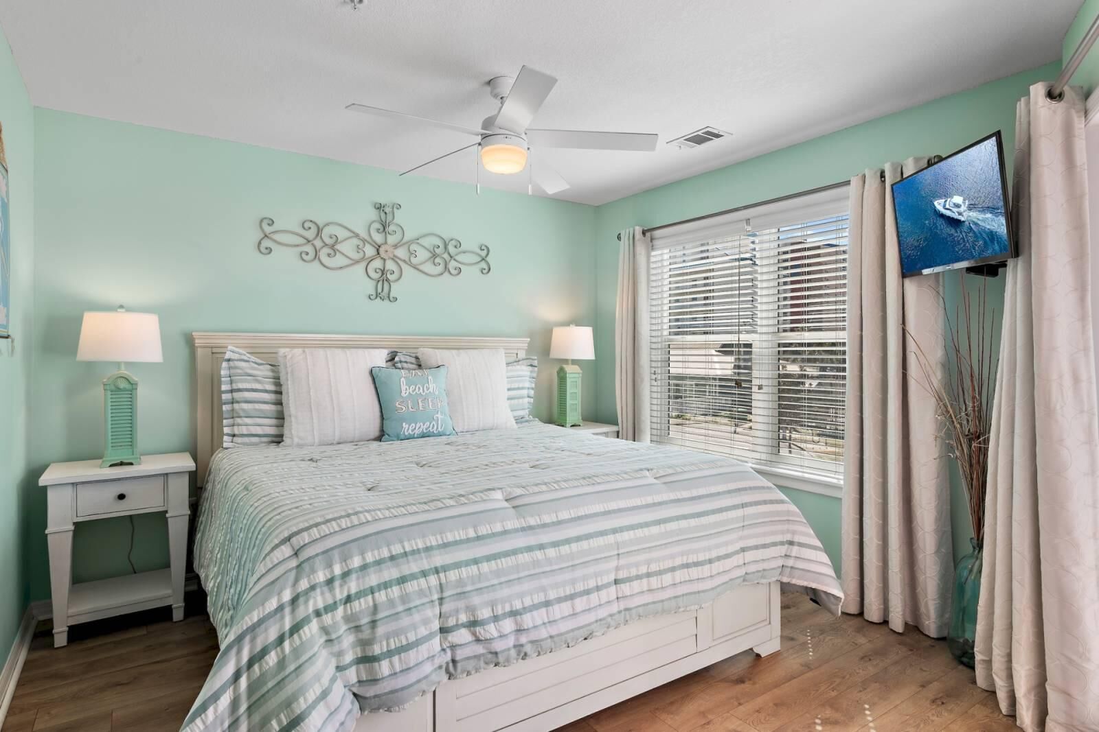 Une chambre à coucher comprenant un lit, deux tables de chevet, un ventilateur de plafond, une fenêtre avec des stores et un téléviseur fixé au mur.