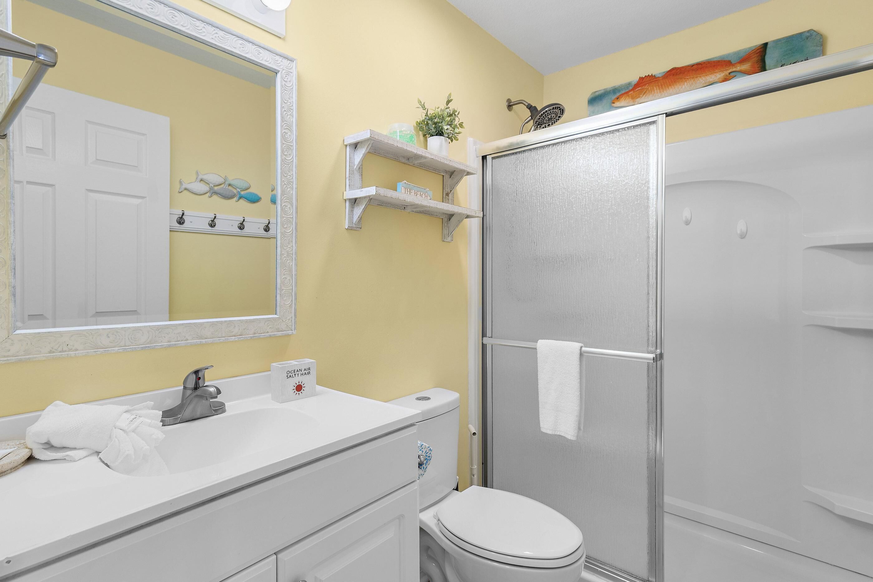 A bathroom with a white sink, toilet, and shower. There are shelves with toiletries and a towel rack.