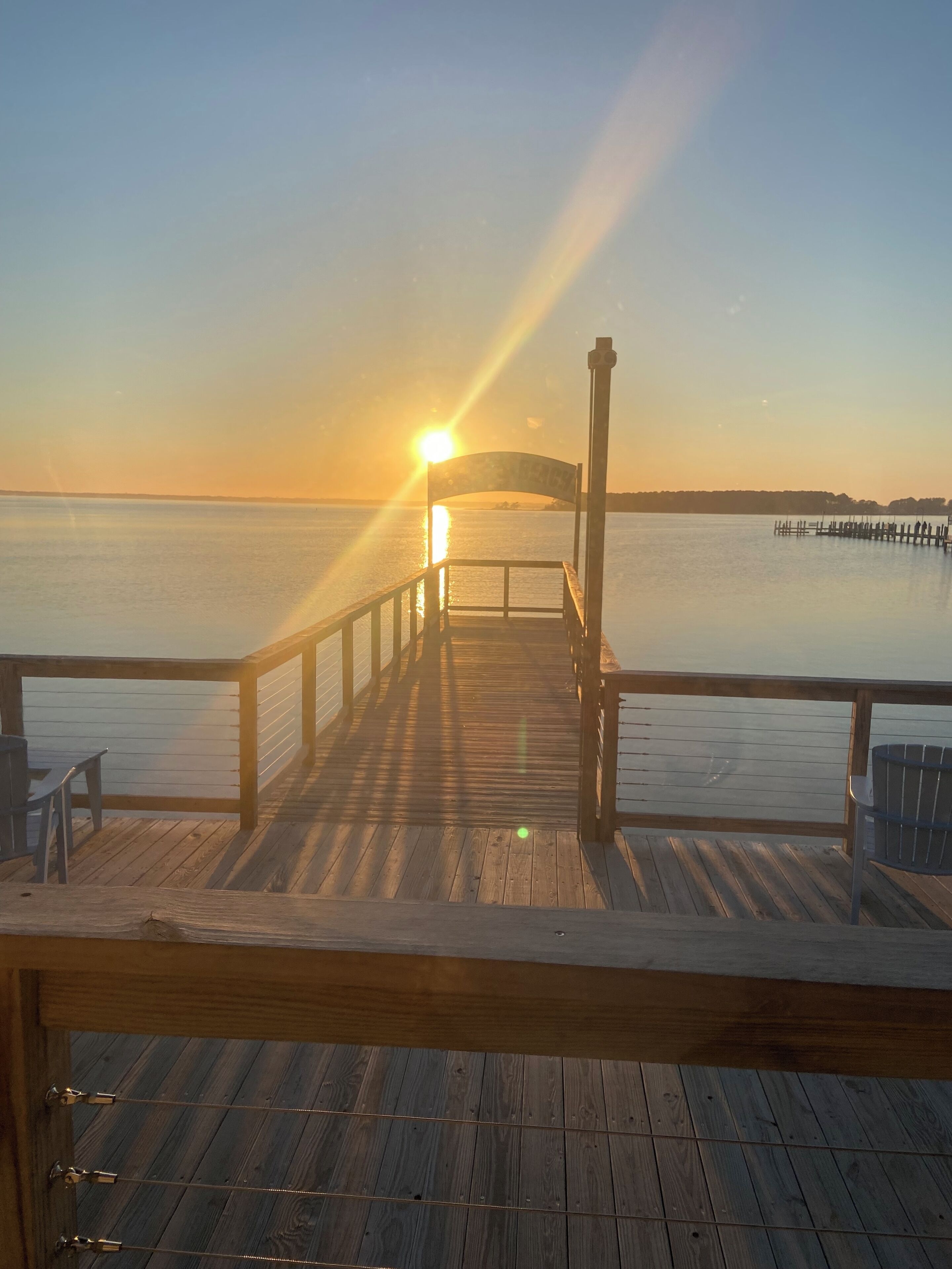 A wooden pier leading to a dock with a railing, overlooking a calm body of water during sunset.