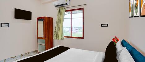 A hotel room with a bed, a wardrobe, a television, an air conditioning unit, a window with curtains, and framed artwork on the wall.