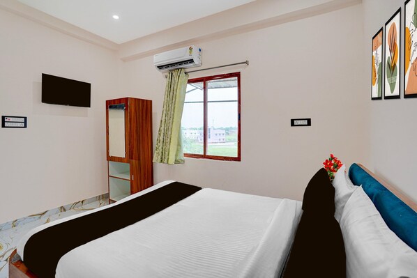 Habitación de hotel con cama, armario, televisor, equipo de aire acondicionado, ventana con cortinas y cuadros en la pared.